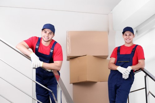 The image depicts two male movers dressed in red shirts, navy blue overalls, white gloves, and navy blue caps, standing on residential staircase landings near the entrance of a house. They are posed in front of a stack of brown cardboard moving boxes, with one on a lower step and two larger boxes behind them, stacked securely. The movers are smiling and appear ready for a house removal task. The interior setting features white walls and a staircase with metal railings, typical of a residential property. Natural light illuminates the scene, highlighting the textures of the cardboard boxes and the fabric of their clothing. This scene is indicative of professional house removals or moving services, with the movers prepared to facilitate the safe transportation of household items, aligning with services offered by Bromley Man with Van in the Bromley area, close to postcode BR1 or BR2. The focus remains on their readiness to assist with moving furniture and boxes in a professional and organized manner within a domestic environment.