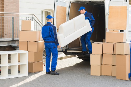 Two removal men dressed in blue uniforms and matching caps are engaged in loading a white sofa into the back of a white moving van parked on a residential street, near a house with beige brick walls and metal railings. The man on the right is holding the sofa upright, carefully guiding it into the van, while the other man stands nearby observing. Surrounding the van are several neatly stacked cardboard boxes of varying sizes, some on a white shelving unit and others directly on the ground, indicating an organized household move. The scene appears to be outdoors during daylight hours, with natural lighting illuminating the workspace, emphasizing the textured fabric of the sofa, the smooth cardboard surfaces of the boxes, and the practical, durable clothing of the movers. This image illustrates a typical house removal process in Bromley, showing the careful handling of furniture and the preparation involved in residential relocations facilitated by Bromley Man with Van, specialists in local removals across the Bromley area.