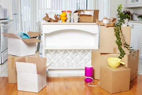 The image depicts a cluttered kitchen area with an assortment of moving boxes and household items scattered across a wooden floor. Several cardboard boxes, some open and partially taped, are positioned in the foreground and background, containing items such as crumpled brown paper, a yellow ceramic bowl, and a small pink portable speaker. A white kitchen island or breakfast bar is visible in the center, with more boxes placed on top, including a cardboard box with brown packing paper and a white mug. The background shows a modern kitchen with white cabinetry, a countertop, and a window with sheer white curtains allowing natural light to fill the space. A leafy green plant trails down from a box on the right, and a pair of brown shoes are seen on top of one of the boxes. The scene appears to capture a house in the process of being packed or prepared for a move, reflecting the services offered by Bromley Man with Van in Bromley, known for residential removals within the local area.