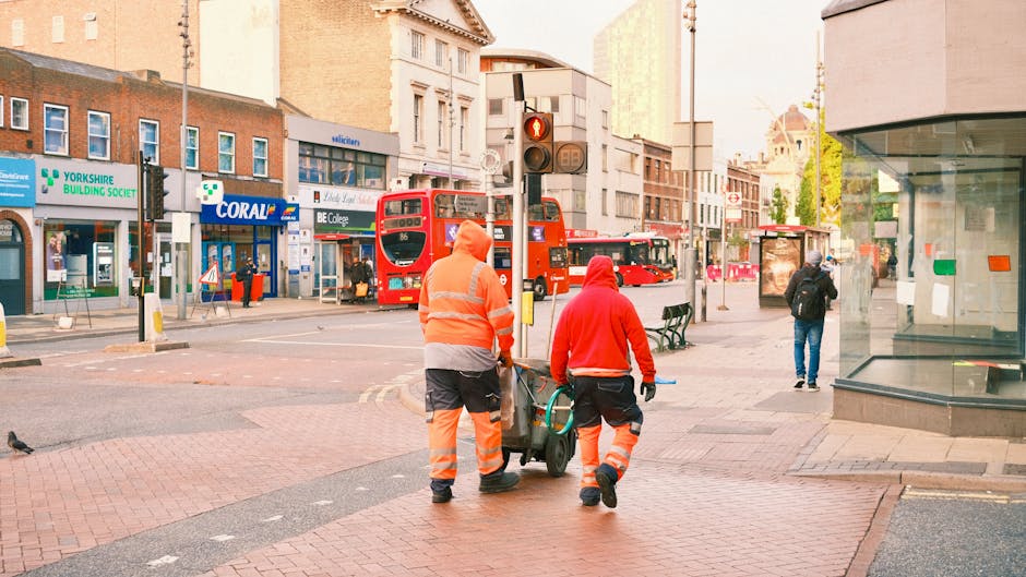 Two removal service workers, dressed in high-visibility orange and red jackets with reflective strips, are loading a wheeled trolley with wrapped furniture and boxes onto a pavement outside a building