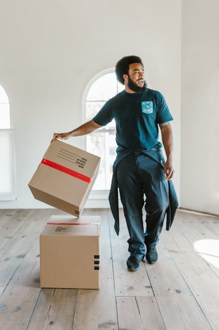 Two movers from Bromley Man With Van, dressed in casual clothing, are inside a bright room with arched windows, engaged in a home relocation process. One mover, with curly hair and a beard, is standin